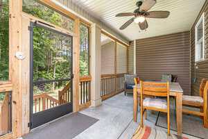 A screened-in porch with a ceiling fan, wood-framed screen door, brown siding, outdoor rug, and a dining table with four chairs. Stairs outside lead to a grassy yard with trees in the background.