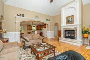 A spacious living room with tan walls, wood floors, a fireplace with a decorative mantel, beige sofas, a glass coffee table, and a view into a kitchen through an arched opening. A large plant stands by the fireplace.
