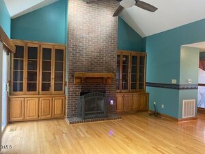 Living room with a brick fireplace centered between two built-in wooden cabinets with glass doors. The room has light wood floors, teal walls, high ceilings, and a ceiling fan. There is a vent and a thermostat on the right wall.