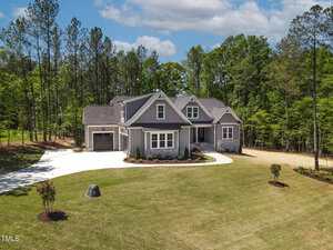 A modern gray house with a front porch and attached garage sits on a large, well-kept lawn surrounded by tall trees. A curved driveway leads to the house, and small shrubs are planted near the entrance.