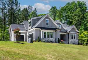 A modern gray house with white trim sits on a sloped lawn. The home has multiple gables, stone accents, a front porch, and is surrounded by trees and landscaped shrubs.