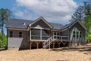 A modern, single-story house with gray siding, multiple gables, a screened-in porch, deck with stairs, and large windows. The yard is covered with straw mulch and surrounded by trees under a partly cloudy sky.
