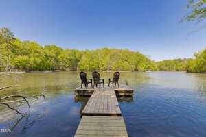 A wooden dock extends over a calm lake, with four black chairs arranged at the end. The lake is surrounded by green trees under a clear blue sky.