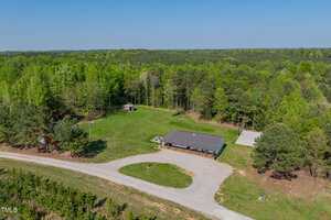 Aerial view of a single-story house with a dark roof, surrounded by grass and trees. A gravel driveway curves in front of the house, and a small shed sits nearby. Dense forest extends into the background.