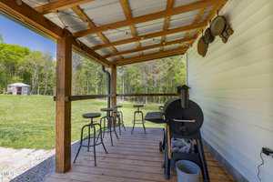A covered wooden deck with a metal grill, three bar stools, and a small counter overlooking a grassy yard. Metal pots and pans hang on the house's white siding. Trees and a shed are visible in the background.