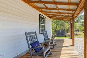 A wooden porch with two gray rocking chairs, a small table, and a swing at the far end. The porch has a wood and metal roof, string lights, and overlooks trees and a gravel area.