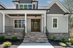 A modern house exterior with gray siding, white trim, a wooden front door with glass panels, two lantern lights, stone steps, metal railings, and small shrubs along a mulch bed.