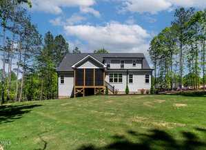A two-story house with light-colored siding and a screened porch sits on a large, grassy yard, surrounded by tall trees under a partly cloudy sky.