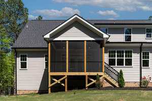 A beige house with white trim features a screened-in porch elevated on wooden posts, accessible by stairs. The lawn is grassy, and several windows are visible along the side of the house. Trees and blue sky are in the background.