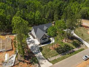 Aerial view of a gray two-story house with a dark roof, surrounded by trees and green lawn, situated on a spacious lot with a paved driveway in a suburban neighborhood under construction.