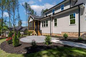 A modern house with gray siding, brick foundation, and a screened porch. The yard features a circular patio, landscaped flower beds, small shrubs, and a concrete walkway. Tall trees and other houses are visible in the background.