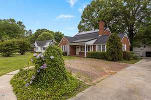 A red brick house with a front porch, chimney, and solar panels on the roof, surrounded by trees and lawn. A vine-covered shrub with purple flowers is in the foreground next to a brick driveway.