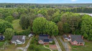 Aerial view of three brick and white houses lined up along a narrow road, surrounded by dense green trees and open grassy areas, with forested land in the background under a blue sky.