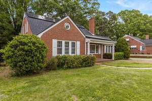 A red brick house with white trim and shutters, solar panels on the roof, a covered porch, and green bushes in front, set on a grassy lawn with another similar house visible in the background.