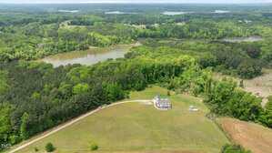 Aerial view of a white house with a driveway, surrounded by open grassy land, trees, and nearby lakes. The landscape features dense forests and waterways extending into the distance.