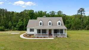 A white two-story house with a gray metal roof, three dormer windows, and a covered front porch sits on a large, grassy lawn with a curved sidewalk and forested trees in the background.