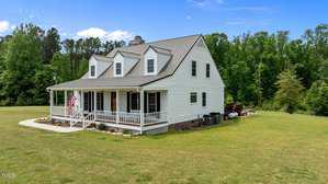 A white two-story house with a metal roof and covered front porch stands on a large grassy lawn, bordered by trees and a wooded area in the background. An American flag hangs near the front steps.