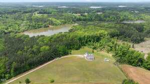 Aerial view of a large house with a driveway on a spacious grassy lot, surrounded by trees, with two lakes and forested areas in the background. The landscape is lush and green under a cloudy sky.