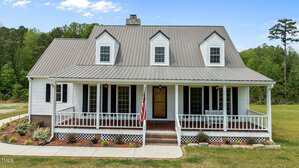 A two-story white house with a metal roof, dormer windows, a covered front porch, and an American flag. The house is surrounded by grass, shrubs, and trees in the background.