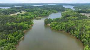 Aerial view of a winding lake surrounded by dense green forest, with a few buildings visible on the left side and open water extending into the background. The scene appears calm and natural.
