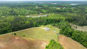 Aerial view of a rural landscape with a single house in the center of a large grassy field, surrounded by dense green trees, a winding road, a lake, and several patches of farmland.