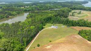 Aerial view of a white house with a driveway, surrounded by green fields, trees, and a river or lake. Farmland and wooded areas are visible, with water in the background.
