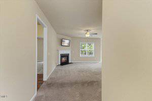 Carpeted living room with beige walls, a ceiling fan with light, a window, a fireplace with a TV above it, and entrances to adjacent rooms. The space is empty and well-lit with natural light.