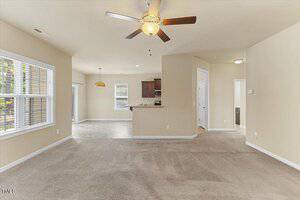 A spacious living room with beige carpet, ceiling fan, and neutral walls, opening to a tiled dining area and kitchen with brown cabinets, stainless steel appliances, and several large windows for natural light.
