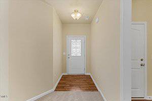 A hallway with beige walls and a ceiling light leads to a white door with a decorative glass panel. The floor transitions from carpet to wood near the door. A second white door with silver handles is partially visible on the right.