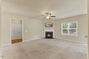 A spacious living room with beige carpet, light-colored walls, a ceiling fan with a light, a white fireplace with a painting above it, and a large window letting in natural light. Double doors lead to another room.
