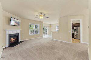 A spacious living room with beige walls and carpet, a ceiling fan, a fireplace with a TV above it, and large windows. The room opens to a tiled dining area and kitchen with stainless steel appliances.