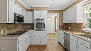 A kitchen with white cabinets, stainless steel appliances, a built-in oven, electric cooktop, double sink, brown tile backsplash, and a window above the sink. Brown tile flooring and tan walls are also visible.