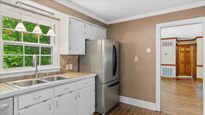 A kitchen with white cabinets, a double sink under a window, stainless steel refrigerator, tiled countertops, and brown tile flooring. The walls are tan, and there is an open doorway to another room.