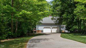 A curved concrete driveway leads to a white house with two garage doors, surrounded by green trees and a well-maintained lawn. The house has a covered porch and brick accents at the base.