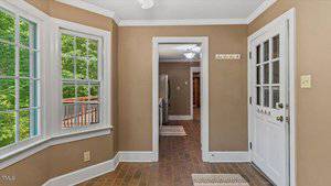A small room with tan walls, white trim, brick flooring, and large windows. A white door with glass panes leads outside. The view extends into a kitchen with visible appliances and a light fixture.