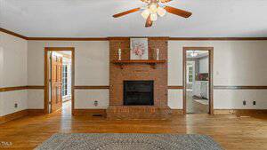 A living room with hardwood floors, a brick fireplace under a wood mantel, a ceiling fan with lights, and two doorways on either side of the fireplace leading to other rooms. A gray patterned rug is on the floor.