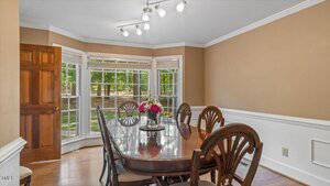 A dining room with a polished wooden table, six chairs, a floral centerpiece, large bay windows showing a green yard, beige walls, white trim, and track lighting on the ceiling.