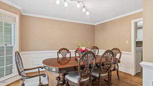 A dining room with tan walls, white trim, a wooden table with six matching chairs, a floral centerpiece, hardwood flooring, a window on the left, and a door opening to a kitchen area on the right.