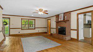 A spacious living room with light-colored walls, wood trim, hardwood floors, a ceiling fan with lights, a brick fireplace, and a large area rug. There are windows, a door, and entryways to other rooms visible.