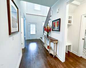 A hallway with dark wood floors, white walls, and a staircase with a black railing. A console table with flowers is against the wall, and two framed pictures are hanging. Doors lead to other rooms on the right.