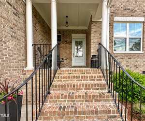 A brick staircase with black metal railings leads up to a covered front porch with white columns. The porch has a white door, brick walls, a barrel planter, and a potted plant on the left side.