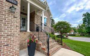 A brick house with a covered front porch, white columns, and black metal railings on the steps. A black planter with pink flowers sits by the stairs. The yard is landscaped with bushes and grass.