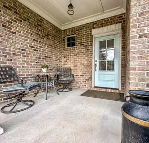 A covered porch with a light blue door, two metal chairs with cushions, a small table with a potted plant, and a large black decorative container on a concrete floor against a brick wall.