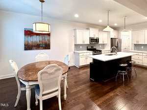 A modern kitchen with white cabinets, stainless steel appliances, a black island with barstools, and a round dining table with four chairs. A painting hangs on the wall near the dining area.