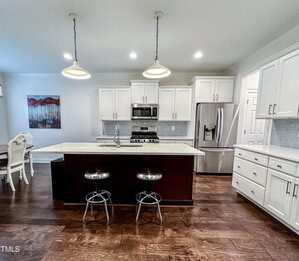 Modern kitchen with white cabinets, stainless steel appliances, a central island with two metal bar stools, dark wood floors, and pendant lights. Dining area with a table and a large painting visible to the left.