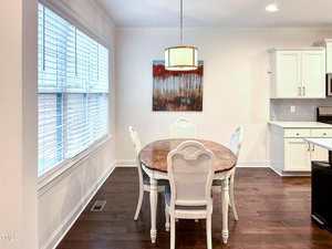 A dining area with a round wooden table and four white chairs on a dark wood floor, a large window with blinds on the left, a kitchen with white cabinets on the right, and a forest-themed painting on the wall.