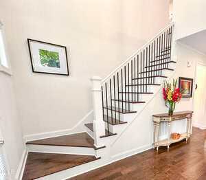 A staircase with dark wooden steps and black railings leads upward along a white wall. A framed picture hangs on the wall, and a small table with a flower arrangement sits beneath the stairs.