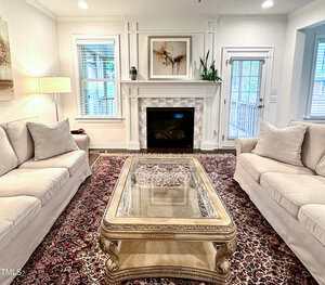 A living room with two cream sofas facing each other, a glass-top ornate coffee table, a fireplace with a white mantel, plants, artwork above the mantel, and a patterned rug on the floor.