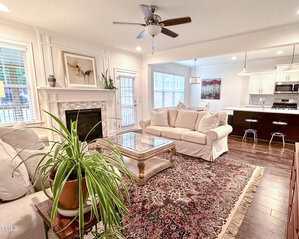 A bright living room with white sofas, a glass coffee table, a fireplace, and a large indoor plant. The open layout leads to a kitchen with white cabinets, a dark island, and three barstools. A patterned rug covers the wood floor.