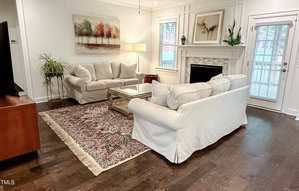 A living room with two white sofas, a glass coffee table, a patterned rug, and a fireplace. There is artwork above the fireplace and sofa, plants, a floor lamp, and windows letting in natural light.